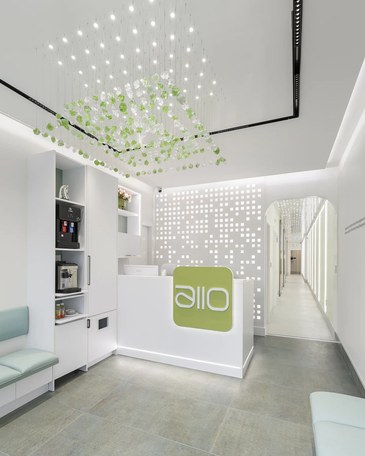 The reception at Allo Dental Clinic: a glowing allo front desk beneath a hanging glass-leaf installation, with the corridor of treatment rooms behind